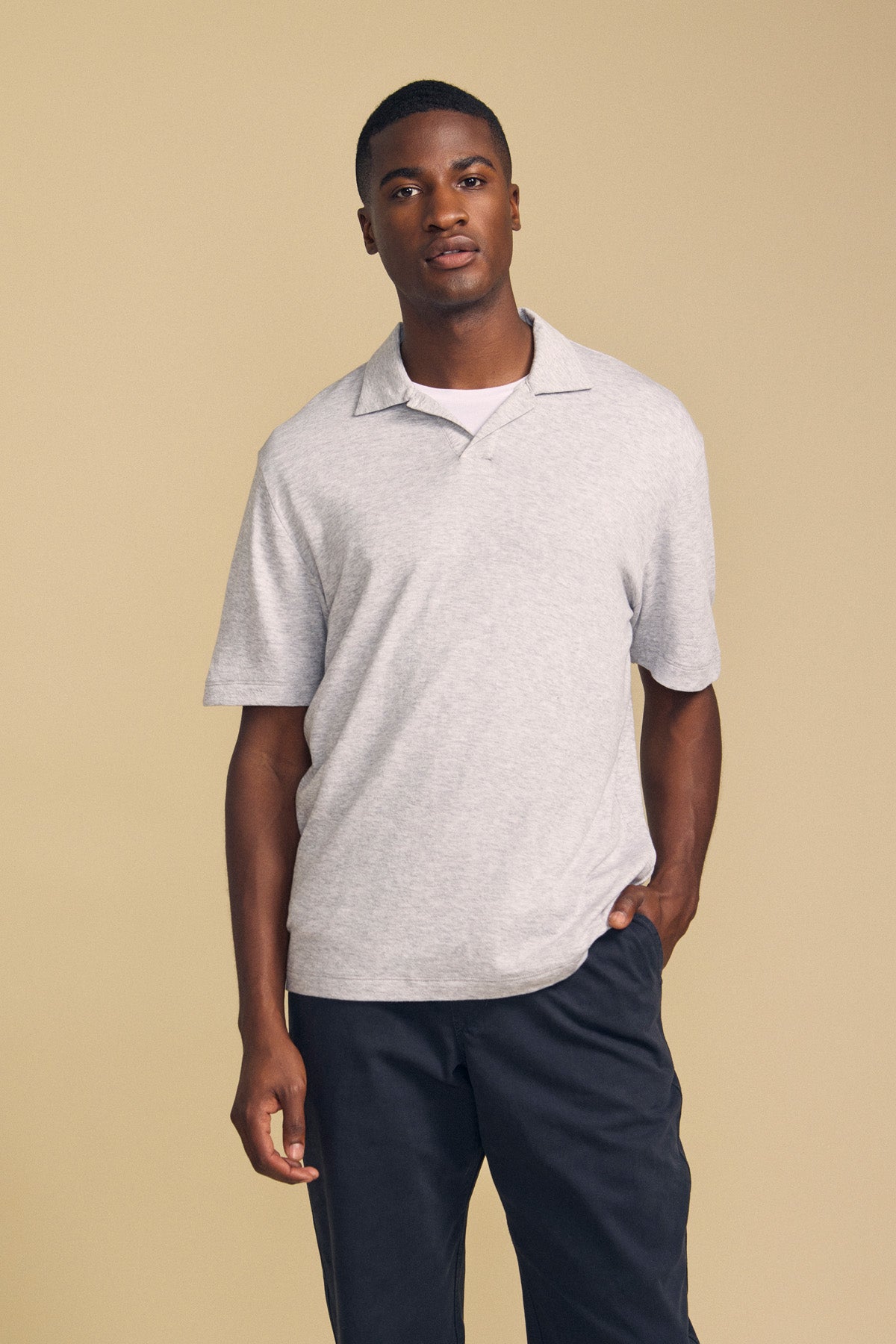 A man stands against a beige background, wearing the Velvet by Graham & Spencer FOX POLO—a light gray short-sleeve polo shirt—layered over a white undershirt and paired with dark pants, with one hand in his pocket.