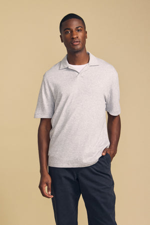 A man stands against a beige background, wearing the Velvet by Graham & Spencer FOX POLO—a light gray short-sleeve polo shirt—layered over a white undershirt and paired with dark pants, with one hand in his pocket.