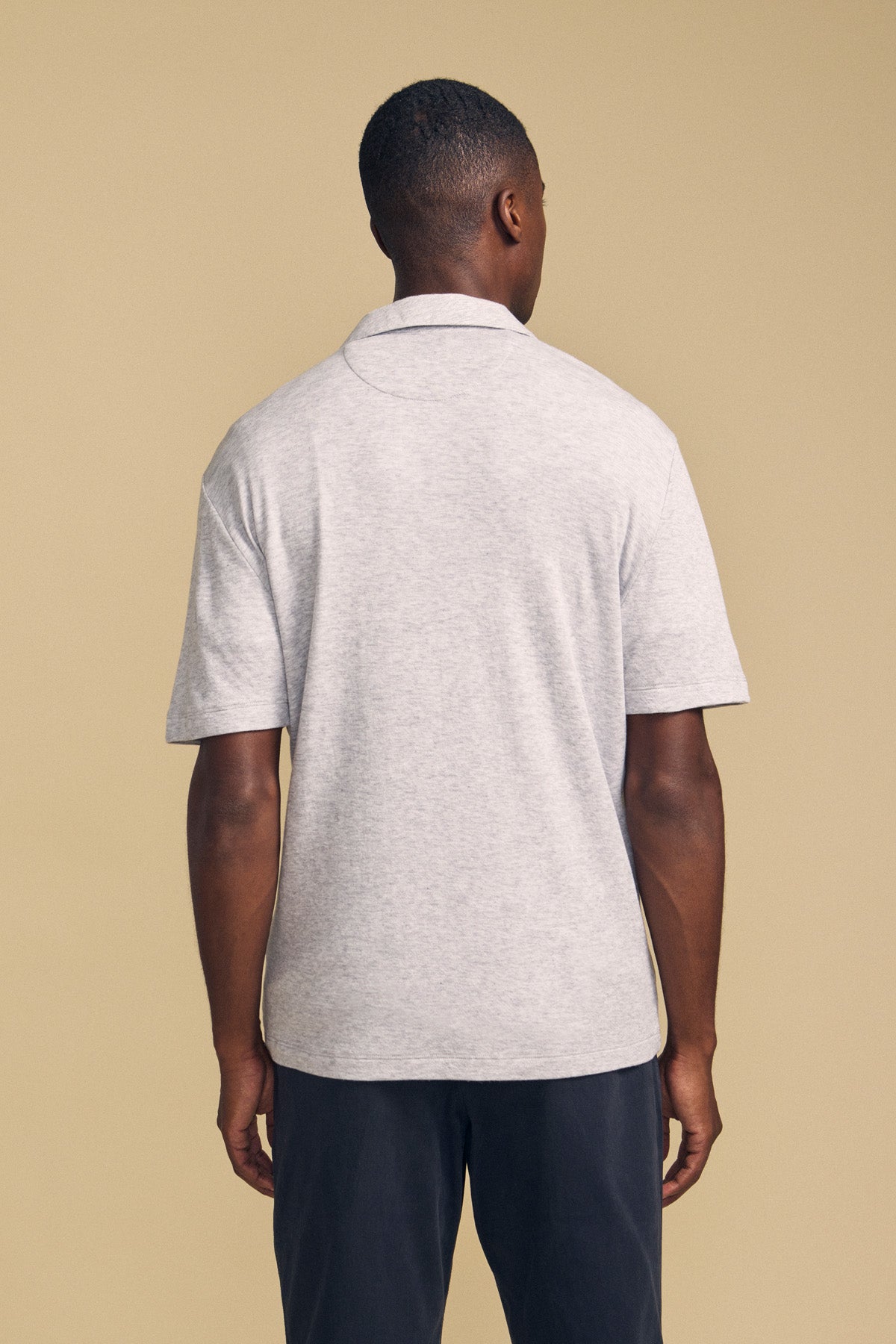 A person stands facing away from the camera, wearing the Velvet by Graham & Spencer FOX POLO in light gray paired with dark pants, set against a plain beige background.