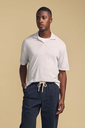 A person stands against a beige background wearing the Velvet by Graham & Spencer FOX POLO, a light gray short-sleeve polo shirt, paired with dark drawstring pants and one hand in their pocket.