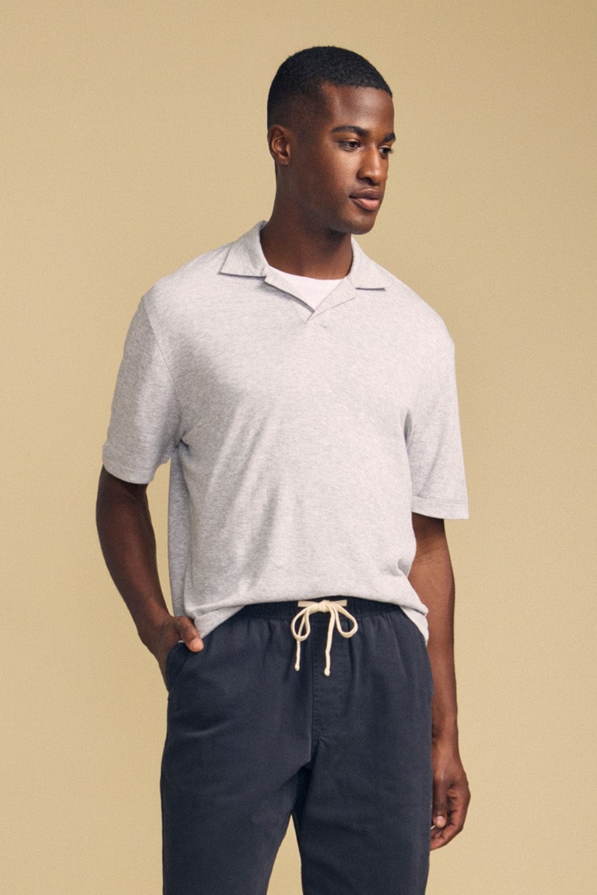 A man wearing the FOX POLO by Velvet by Graham & Spencer, a light gray short-sleeve polo shirt, paired with dark drawstring pants, stands against a beige background looking slightly to the side.