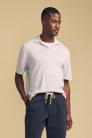 A man wearing the FOX POLO by Velvet by Graham & Spencer, a light gray short-sleeve polo shirt, paired with dark drawstring pants, stands against a beige background looking slightly to the side.