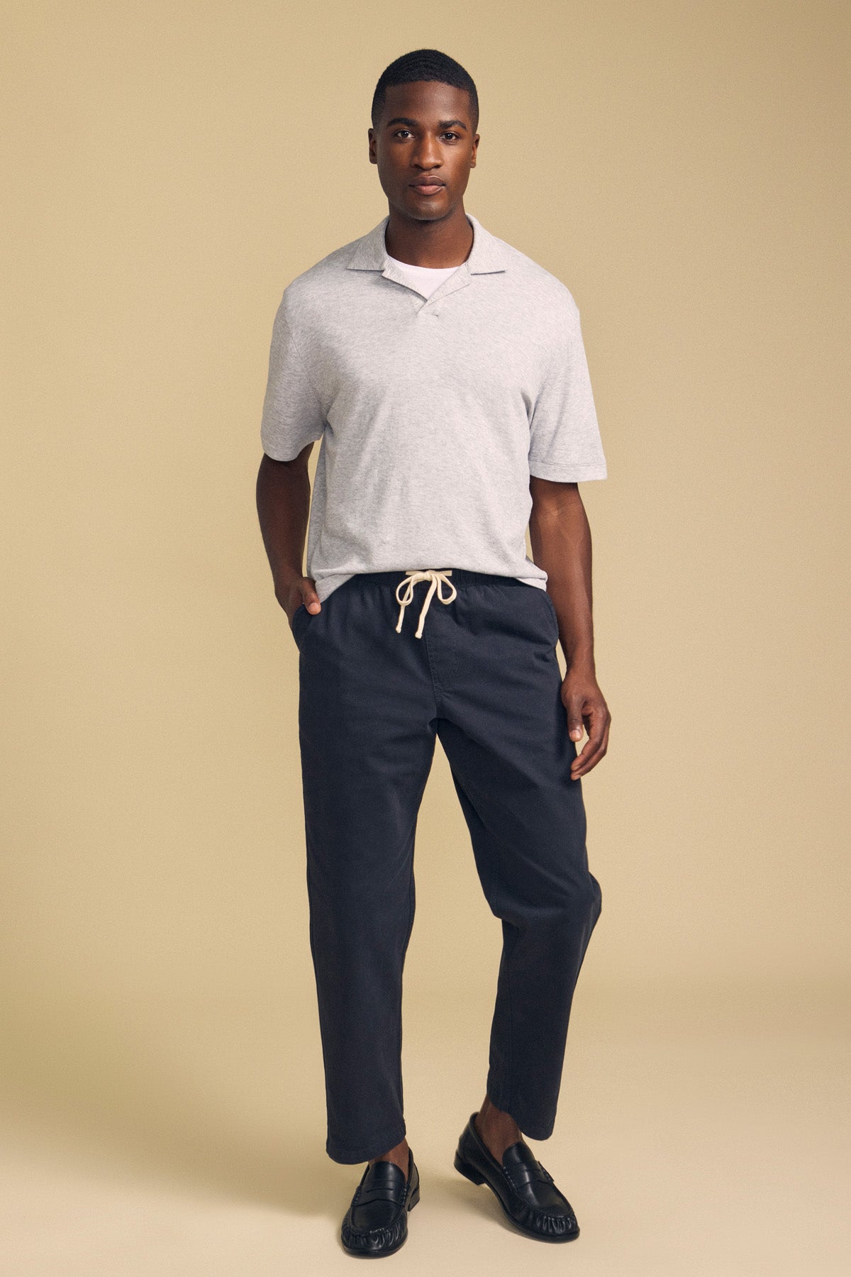 A man stands against a beige background wearing the FOX POLO by Velvet by Graham & Spencer—a light gray polo shirt—paired with dark drawstring pants and black loafers, with one hand in his pocket.
