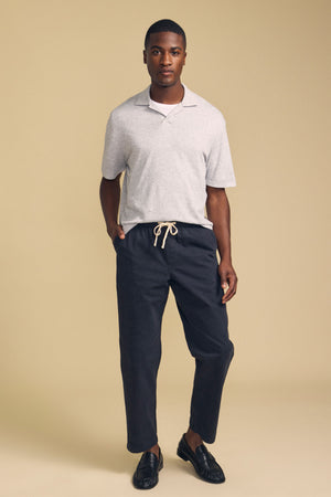 A man stands against a beige background wearing the FOX POLO by Velvet by Graham & Spencer—a light gray polo shirt—paired with dark drawstring pants and black loafers, with one hand in his pocket.