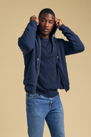 A man wearing a navy blue Velvet by Graham & Spencer CARTER FULL-ZIP HOODIE over a dark sweater and blue jeans, pulling the hood up.