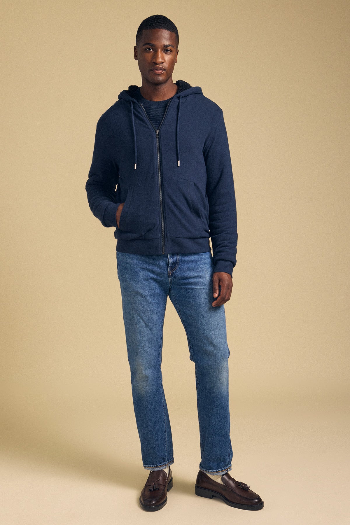 A man wearing a navy blue CARTER FULL-ZIP HOODIE over a dark shirt, paired with blue jeans and brown loafers.-46288221569217