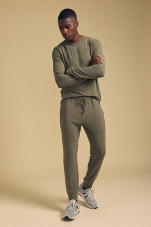 A man stands with arms crossed, wearing the ELLIS SWEATPANT by Velvet by Graham & Spencer—relaxed fit jogger sweatpants in olive green soft cotton jersey—paired with a matching long-sleeve shirt and gray sneakers, set against a beige background.