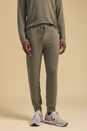 A person wearing the ELLIS SWEATPANT by Velvet by Graham & Spencer in olive green cotton jersey with a relaxed fit, paired with a matching long-sleeve top and gray sneakers, standing against a beige background.