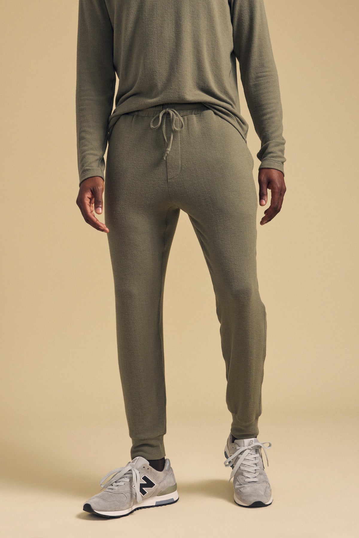 A man stands against a beige background wearing an olive green long-sleeve shirt and the ELLIS SWEATPANT by Velvet by Graham & Spencer, relaxed fit cotton jogger pants with a drawstring, paired with gray New Balance sneakers.-42092470010049