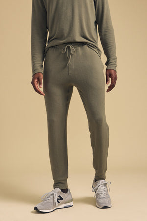 A man stands against a beige background wearing an olive green long-sleeve shirt and the ELLIS SWEATPANT by Velvet by Graham & Spencer, relaxed fit cotton jogger pants with a drawstring, paired with gray New Balance sneakers.