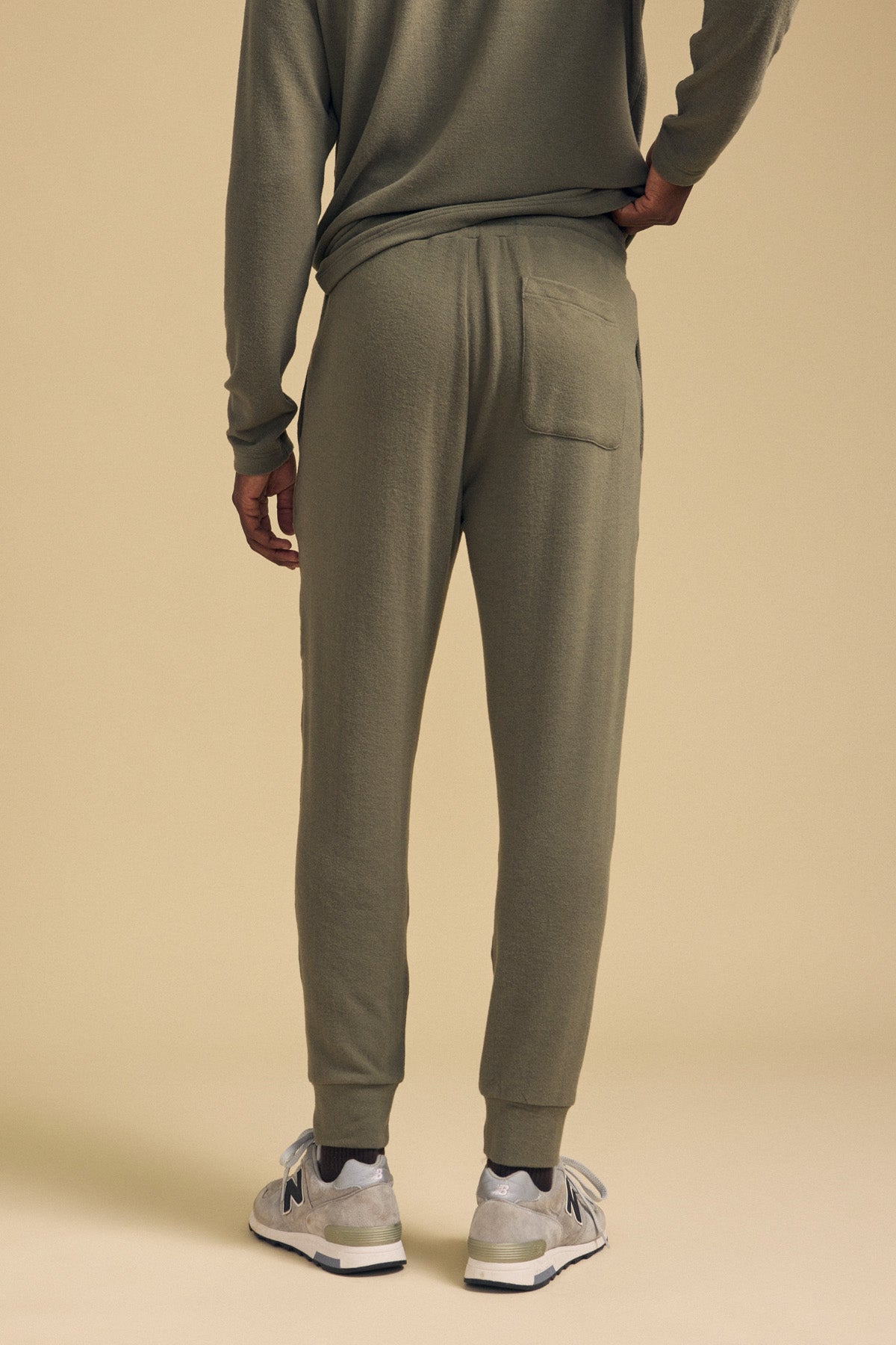 A person stands with their back to the camera, wearing the ELLIS SWEATPANT by Velvet by Graham & Spencer in olive green cotton jersey with a relaxed fit, paired with a matching top and gray sneakers, set against a beige background.-42092470042817