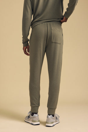 A person stands with their back to the camera, wearing the ELLIS SWEATPANT by Velvet by Graham & Spencer in olive green cotton jersey with a relaxed fit, paired with a matching top and gray sneakers, set against a beige background.