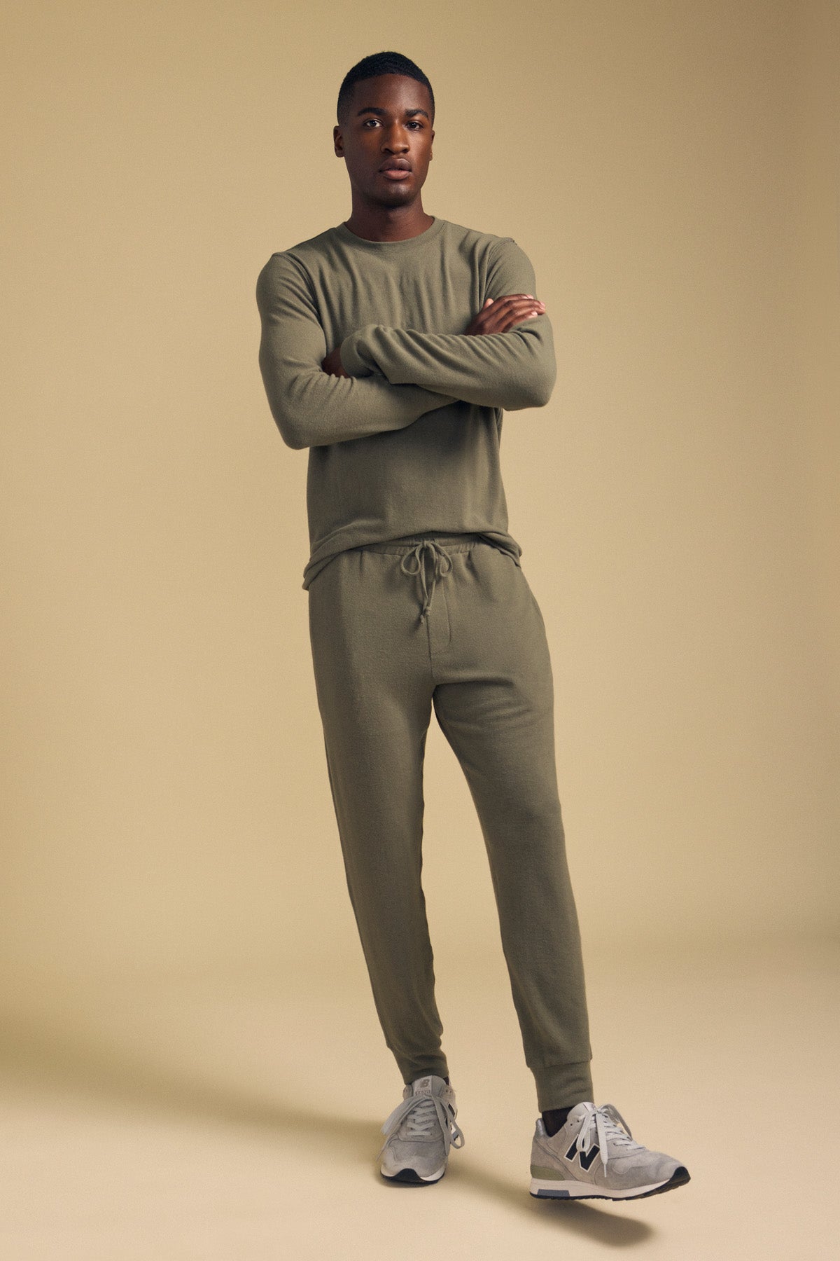A man stands with arms crossed, wearing the Velvet by Graham & Spencer ELLIS SWEATPANT in olive green cotton jersey with a relaxed fit, paired with gray sneakers, against a plain beige background.-42092469813441