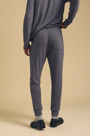 Model wearing the ELLIS SWEATPANT by Velvet by Graham & Spencer in grey, paired with a matching long-sleeve top, stands on a beige background with one hand on her hip and facing away.