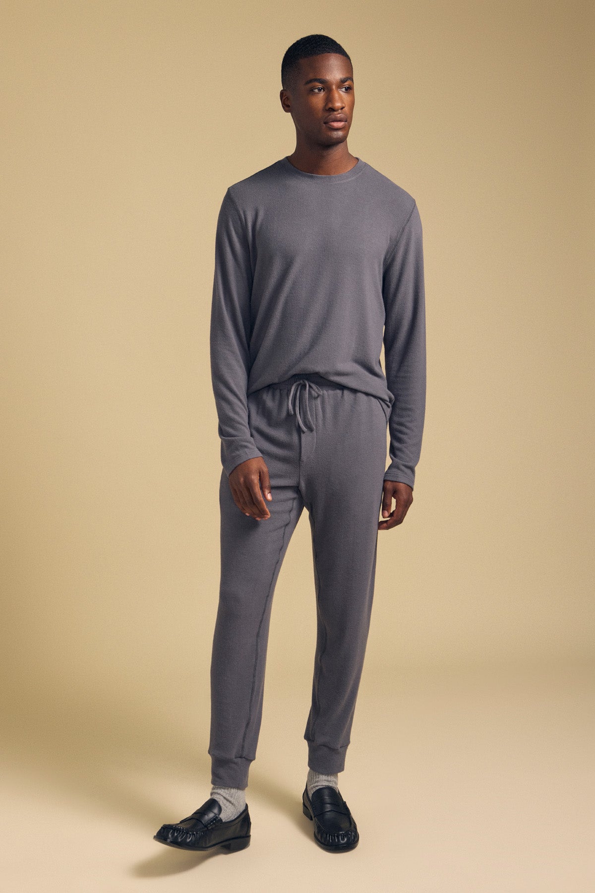 A man stands in a neutral setting wearing the ELLIS SWEATPANT jogger pants by Velvet by Graham & Spencer with a matching long-sleeve gray top, paired with black loafers and light socks.