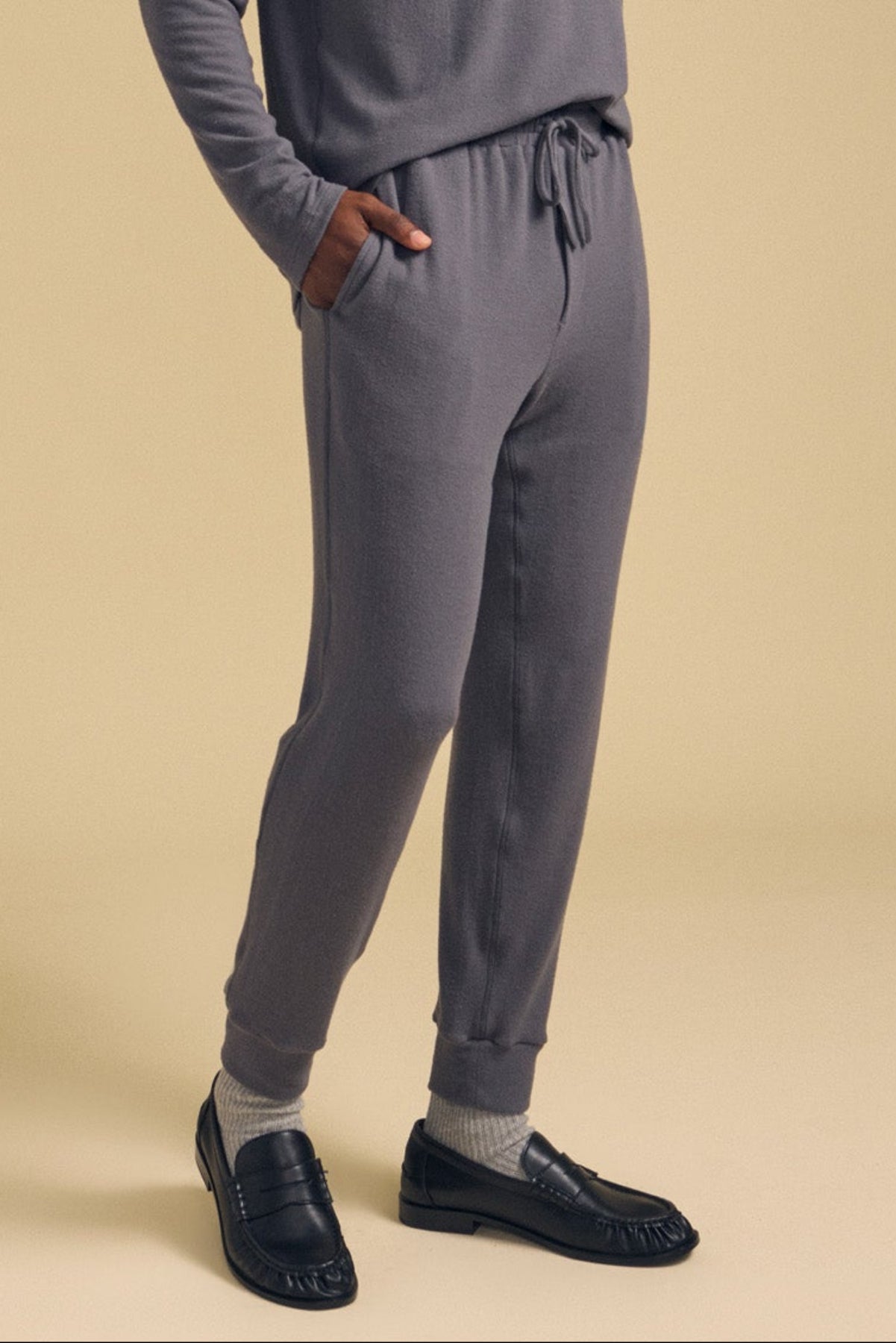 A person wearing the ELLIS SWEATPANT by Velvet by Graham & Spencer, paired with gray socks and black loafers, stands with one hand in their pocket against a beige background.