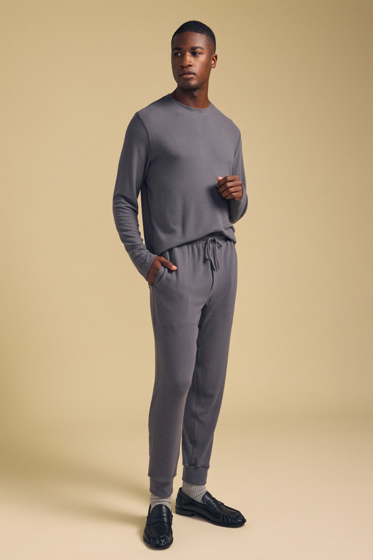 A man stands in a neutral pose against a plain beige background, wearing the matching grey ELLIS SWEATPANT from Velvet by Graham & Spencer, paired with a coordinating long-sleeve top and black shoes.