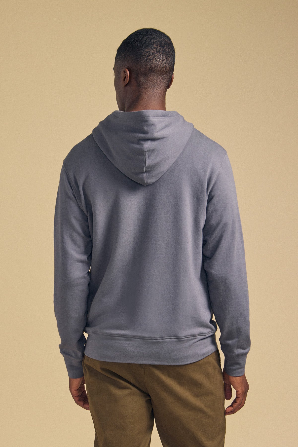 A man stands facing away, wearing the EVERSON HOODIE by Velvet by Graham & Spencer and khaki pants against a plain beige background—a refined staple in the modern wardrobe.-42092470862017