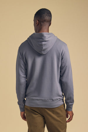 A man stands facing away, wearing the EVERSON HOODIE by Velvet by Graham & Spencer and khaki pants against a plain beige background—a refined staple in the modern wardrobe.