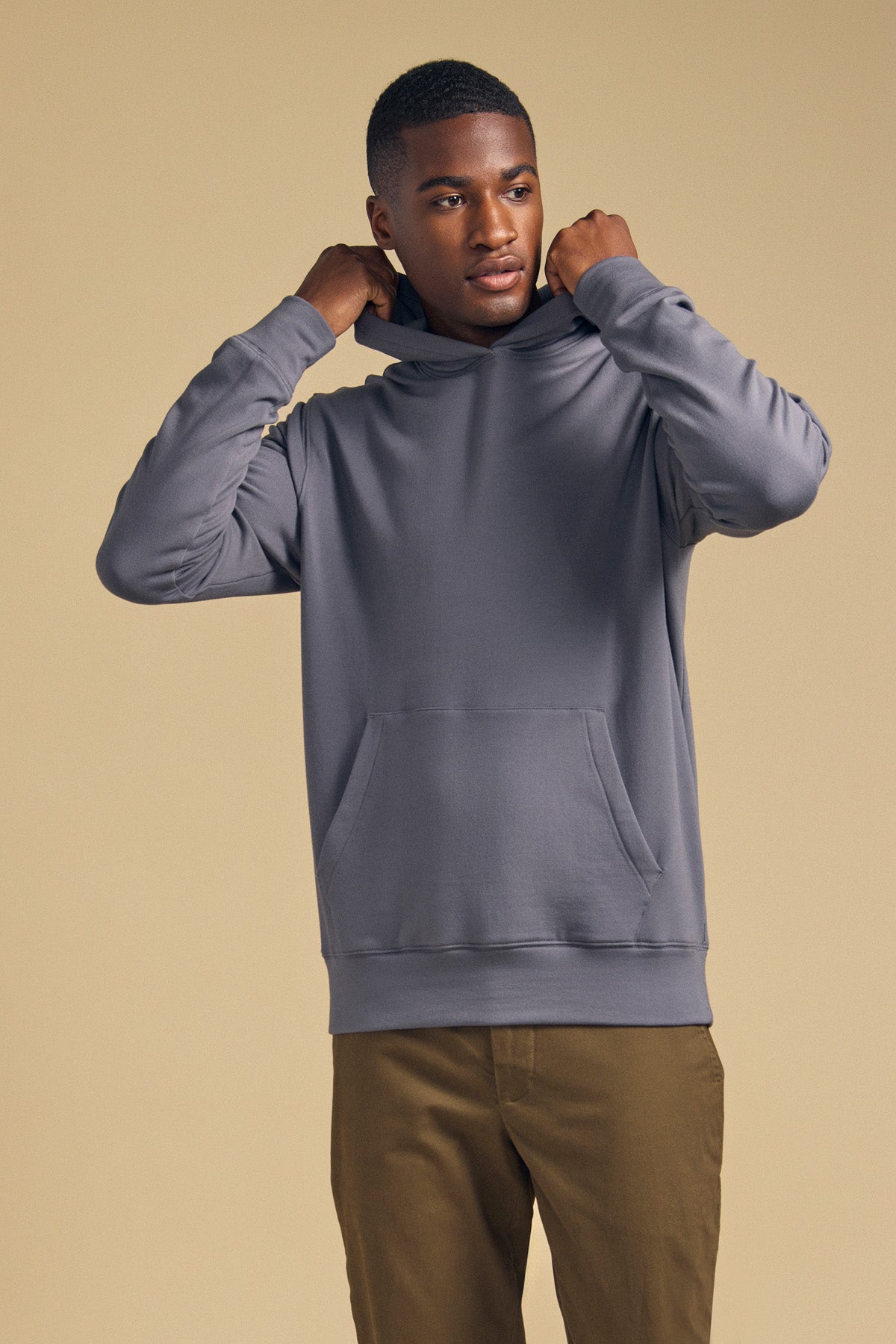 A man wearing the Velvet by Graham & Spencer EVERSON HOODIE and brown pants adjusts his hood while standing against a plain beige background.-42092470665409