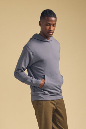 A man stands against a beige background, wearing the EVERSON HOODIE by Velvet by Graham & Spencer—a gray luxe fleece pullover—paired with olive green pants, his hands tucked into the hoodie’s front pocket, making it a perfect addition to any modern wardrobe.