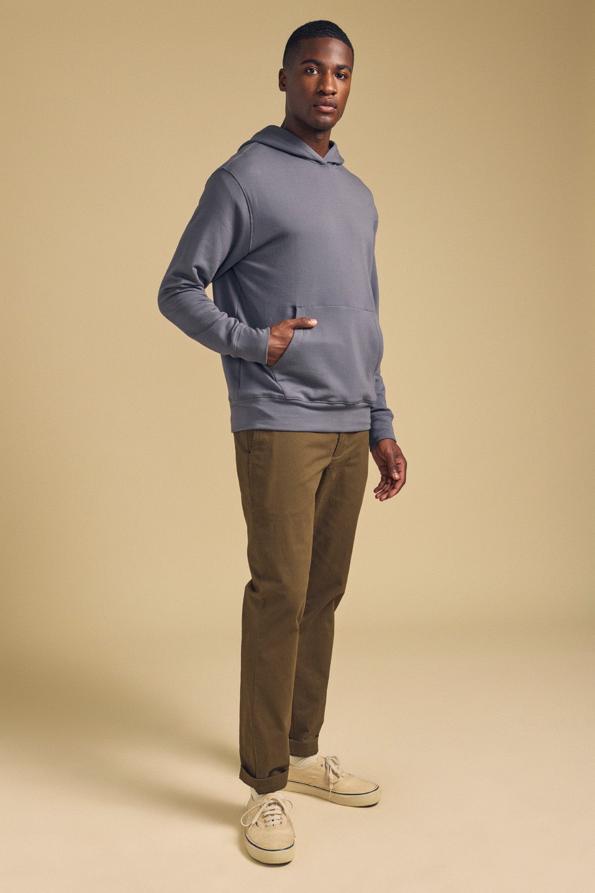 A man stands facing slightly left, wearing the EVERSON HOODIE by Velvet by Graham & Spencer, paired with khaki pants and beige sneakers against a plain beige background—a perfect ensemble for the modern wardrobe.-42092470829249