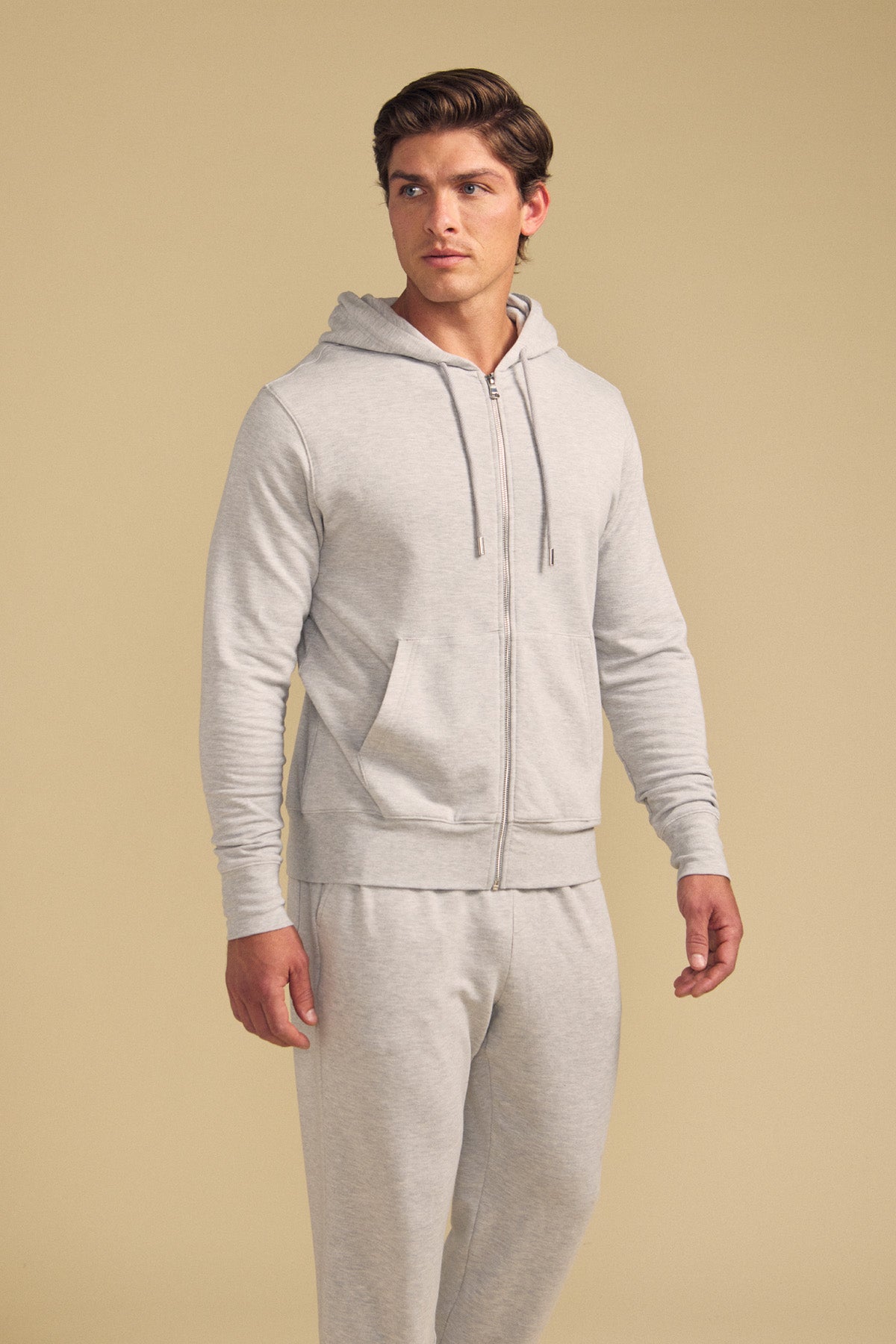 A man wearing the GUS HOODIE by Velvet by Graham & Spencer and matching sweatpants stands against a beige background, looking to the side.-41702021267649