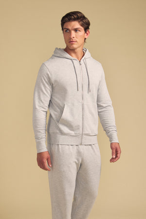 A man wearing the GUS HOODIE by Velvet by Graham & Spencer and matching sweatpants stands against a beige background, looking to the side.