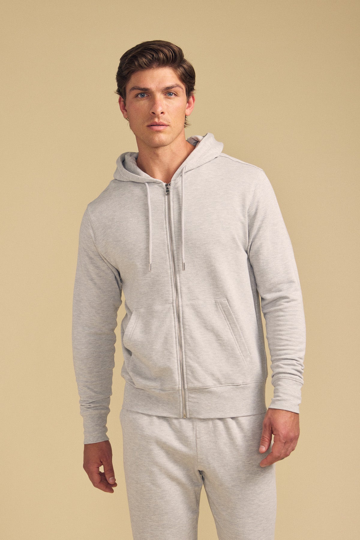 A man wears the GUS HOODIE by Velvet by Graham & Spencer—a light gray zip-up hoodie paired with matching sweatpants—standing against a beige background.
