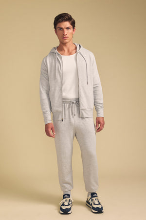 A man stands facing forward, wearing the JACOBY JOGGER by Velvet by Graham & Spencer with a light gray zip-up hoodie, white t-shirt, and navy sneakers against a plain beige background.