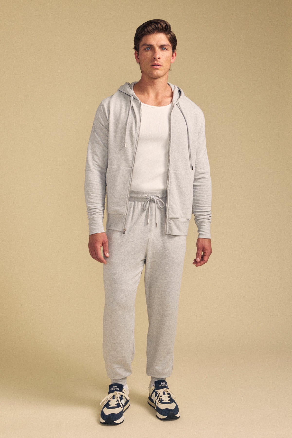 A man faces forward wearing the GUS HOODIE by Velvet by Graham & Spencer in light gray, with matching joggers, a white tee, and blue sneakers against a plain beige background.-41702021103809