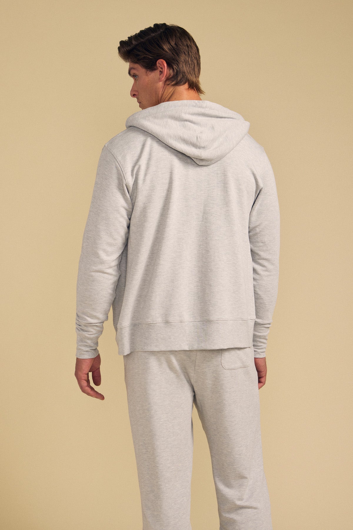 A person stands with their back to the camera, wearing the GUS HOODIE and matching sweatpants by Velvet by Graham & Spencer in light gray, set against a plain beige background.-41702020972737