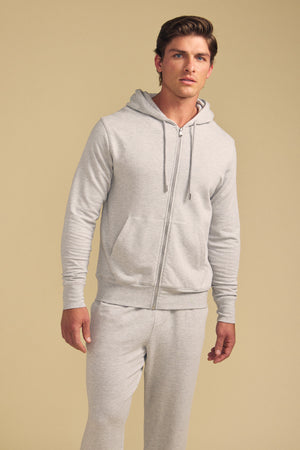 A man stands against a plain beige background wearing the GUS HOODIE by Velvet by Graham & Spencer—a light gray zip-up hoodie—paired with matching sweatpants.