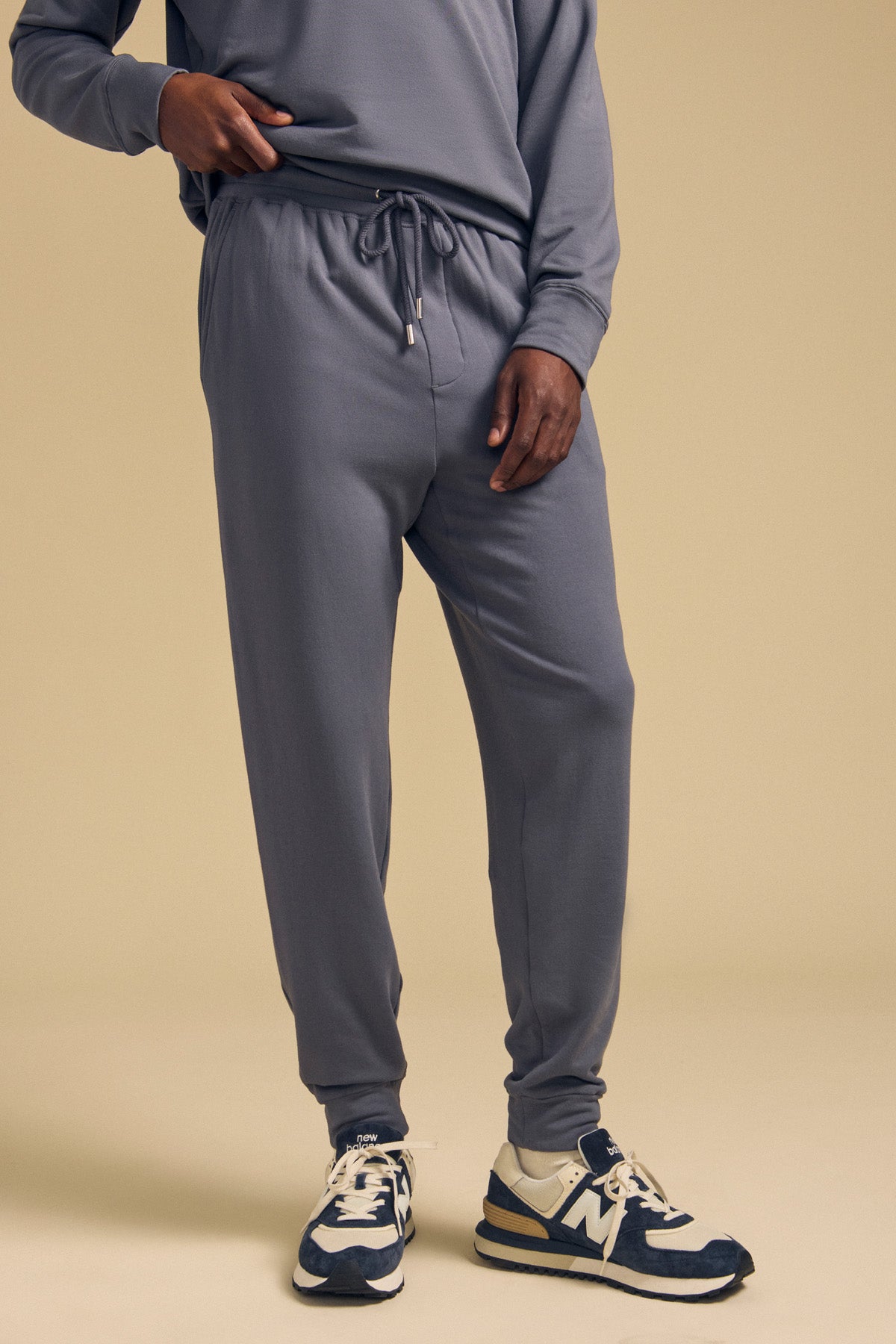 Wearing the JACOBY JOGGER by Velvet by Graham & Spencer—gray jogger pants crafted from a soft modal-cotton-elastane blend—paired with navy-and-white sneakers and set against a beige background, this look embodies everyday comfort.