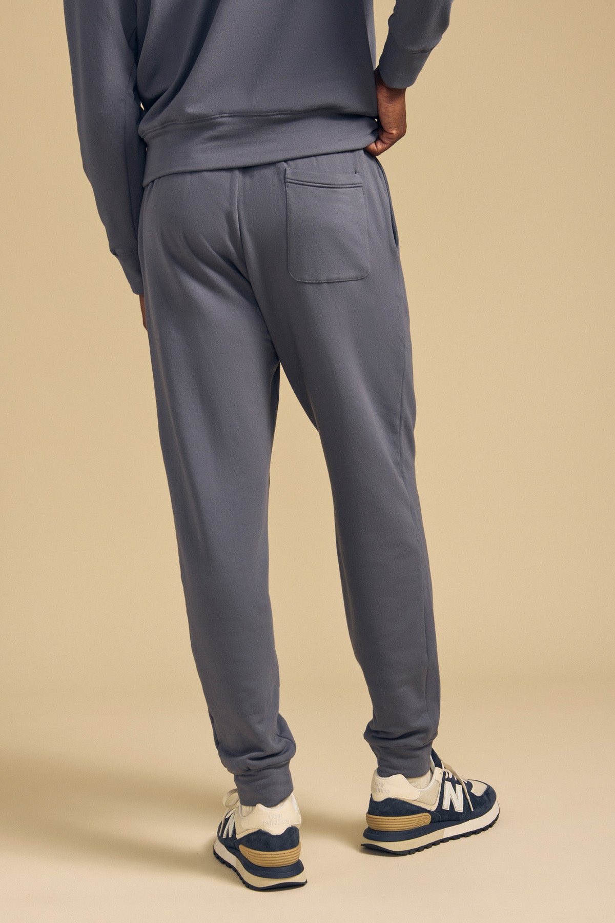 A person stands with their back to the camera, dressed in the JACOBY JOGGER sweatpants by Velvet by Graham & Spencer, with a matching sweatshirt, white socks, and blue sneakers against a beige background—an easygoing look crafted for everyday comfort.-42092477776065