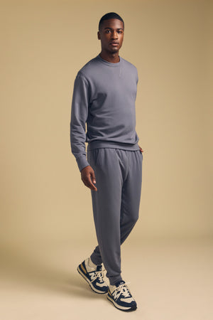 A man stands on a beige background wearing the JACOBY JOGGER sweatshirt and pants by Velvet by Graham & Spencer, crafted from a soft modal-cotton-elastane blend for everyday comfort, paired with black and white sneakers.