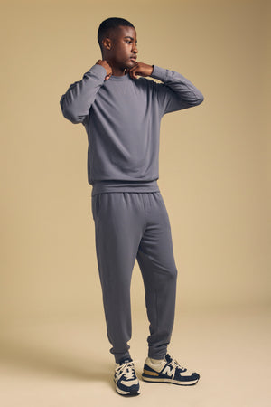 A person models the Velvet by Graham & Spencer JACOBY JOGGER and coordinating grey sweatshirt on a beige background, gazing to the side with hands lifted near their neck.