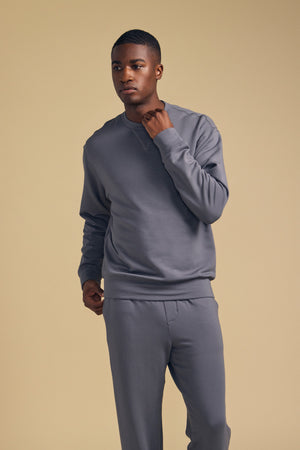 A man wearing the OTIS SWEATSHIRT by Velvet by Graham & Spencer and matching gray luxe fleece pants stands against a plain beige background, one hand touching his neck and the other hanging by his side, showcasing the relaxed fit.