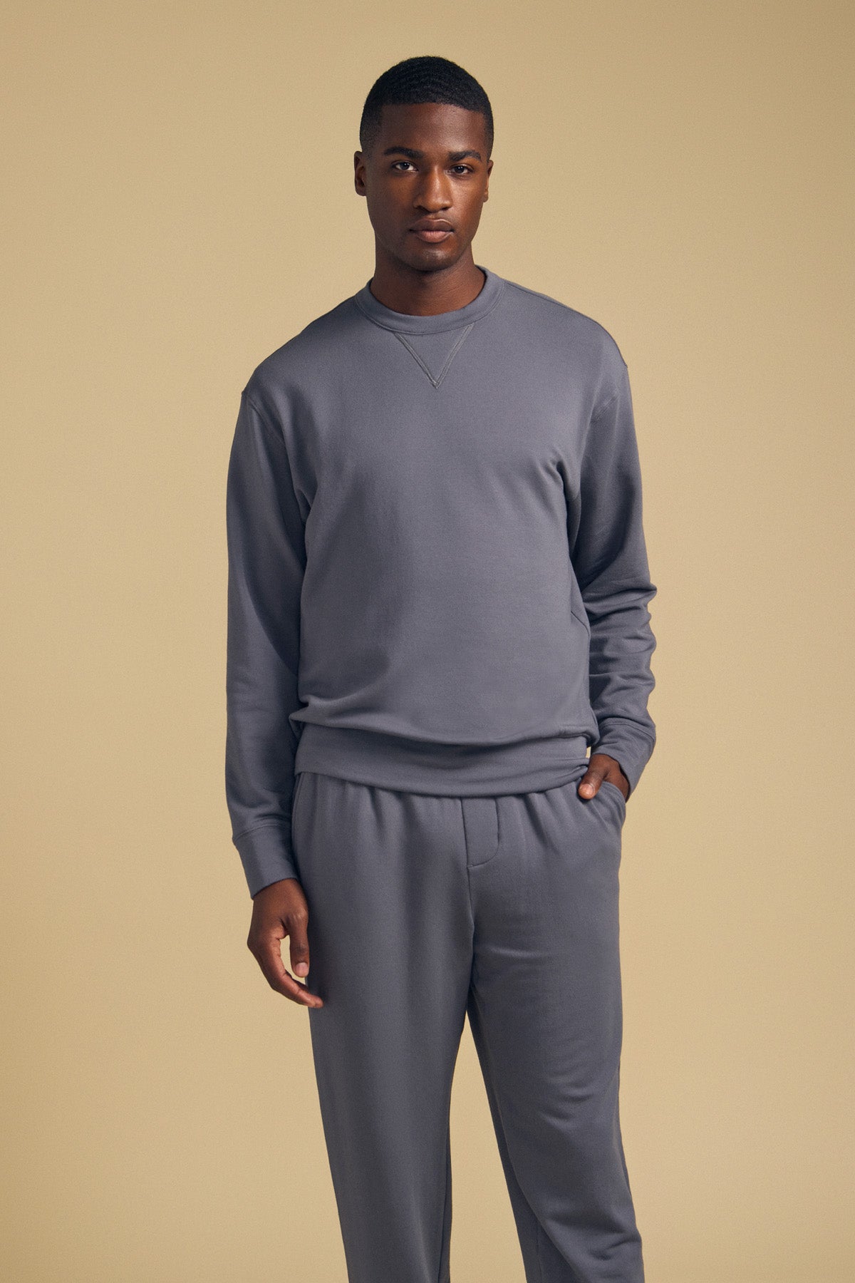 A man stands against a beige background wearing Velvet by Graham & Spencer’s OTIS SWEATSHIRT, paired with matching luxe fleece pants. The relaxed fit and crew neck design are highlighted as he poses with one hand in his pocket and a neutral expression.