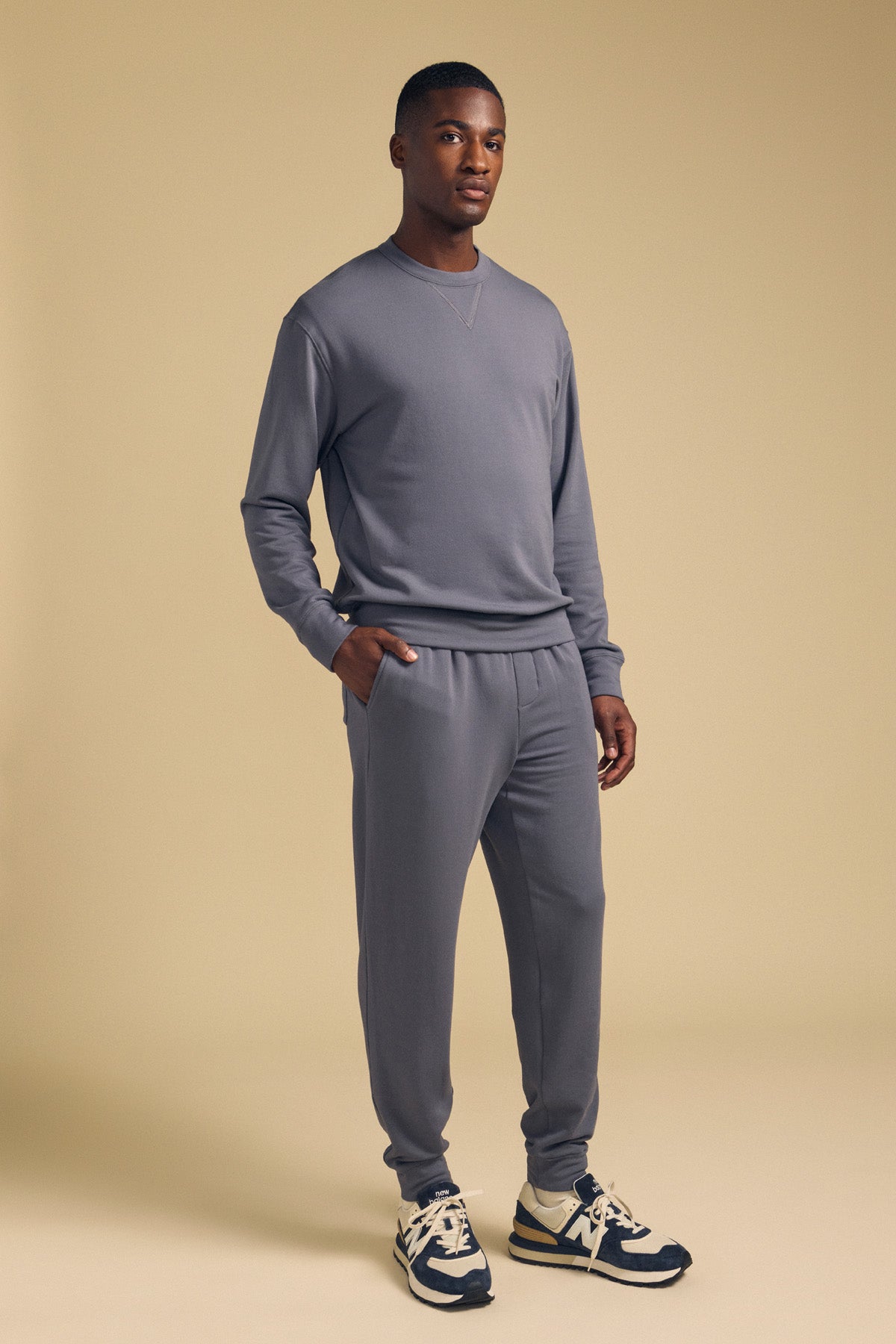 A man stands in a studio wearing the Velvet by Graham & Spencer OTIS SWEATSHIRT in gray paired with matching relaxed-fit joggers, one hand in his pocket, and black and white sneakers.