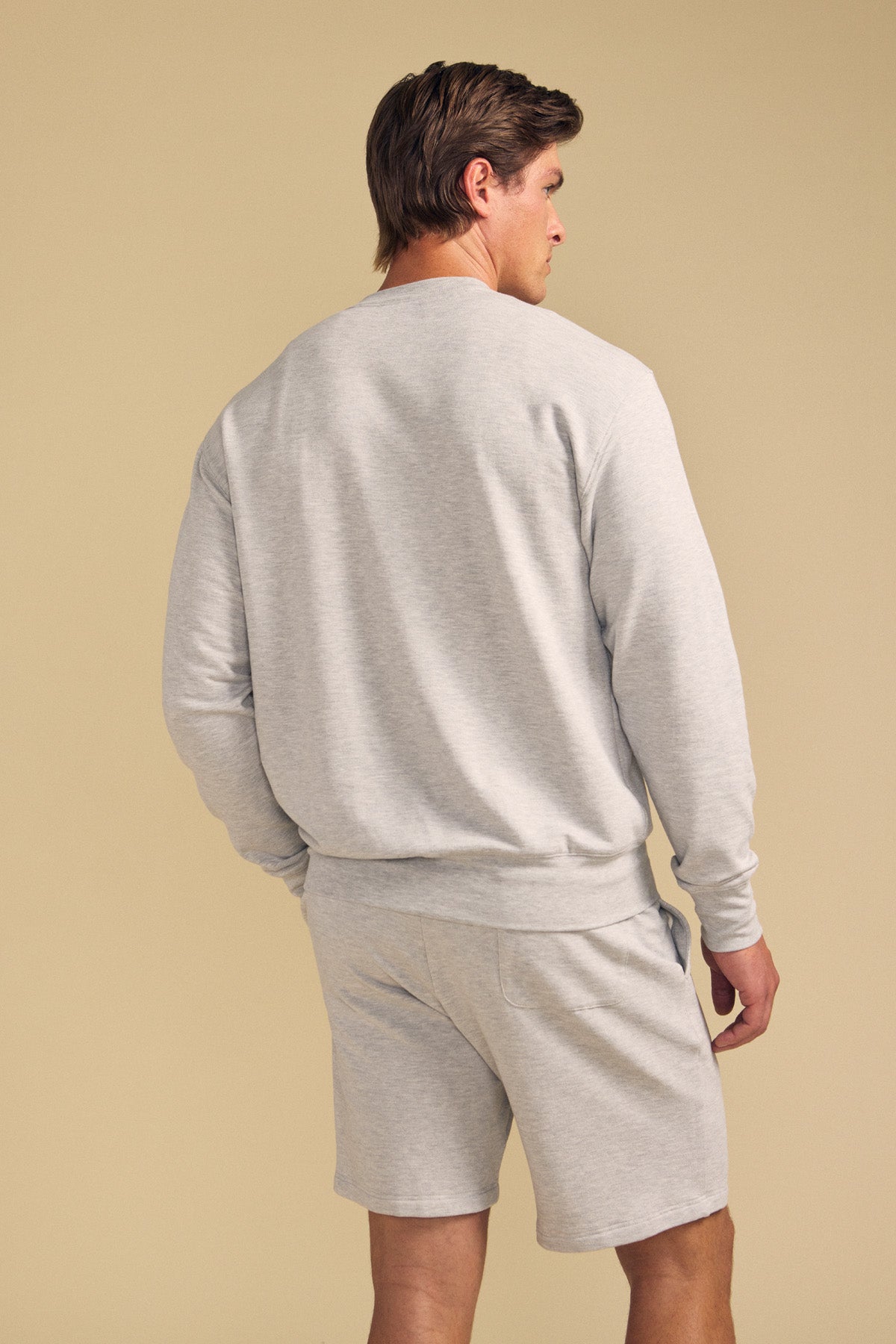 A man faces away, wearing the OTIS SWEATSHIRT by Velvet by Graham & Spencer in light gray with matching shorts, set against a plain beige background.