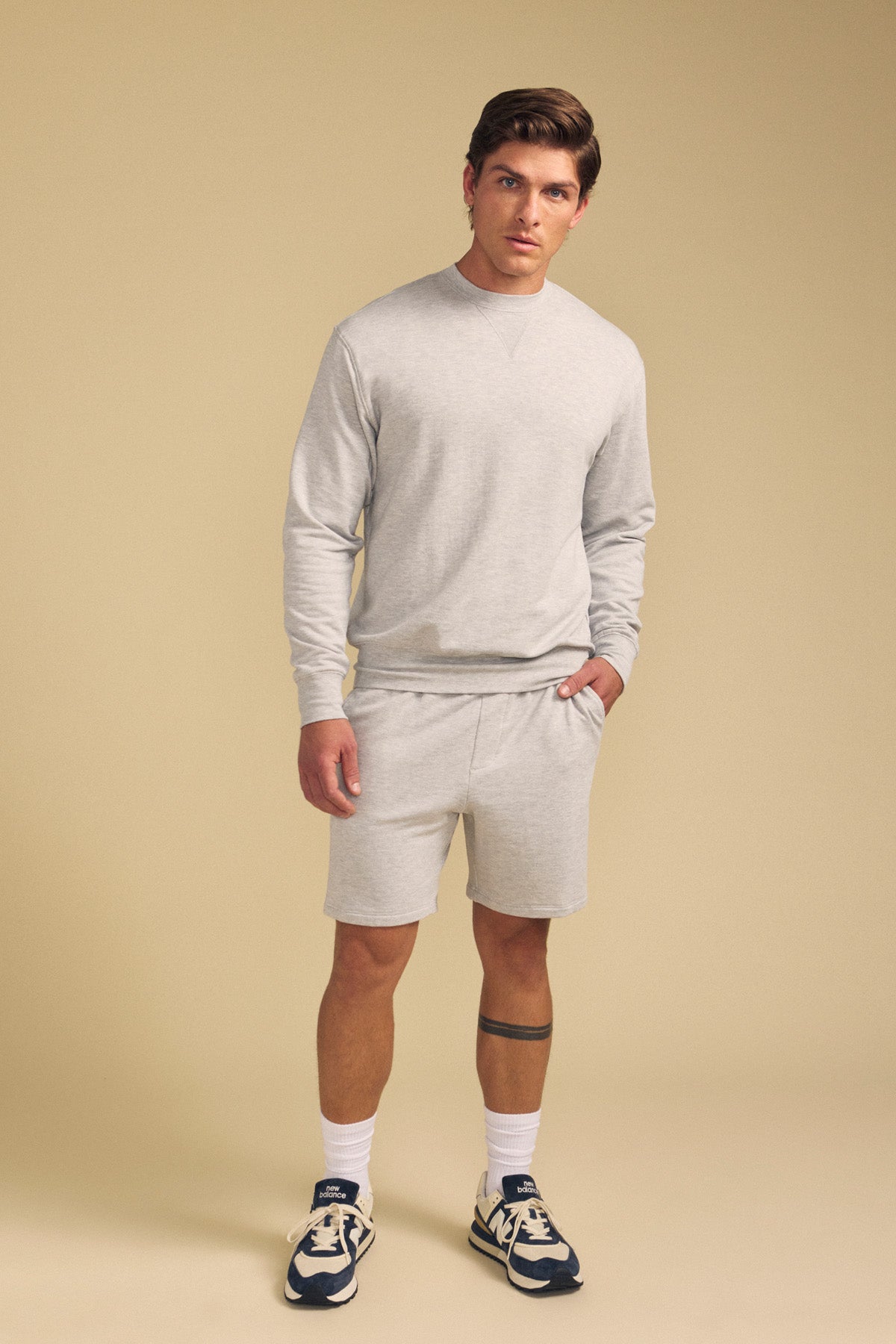 A man stands against a beige background wearing the OTIS SWEATSHIRT by Velvet by Graham & Spencer with matching shorts, white socks, and navy sneakers. One hand is in his pocket.-41702161645761