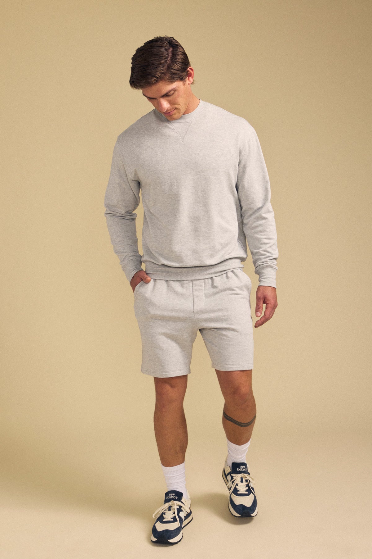 A man stands against a beige background wearing Velvet by Graham & Spencer's QUENTIN SWEATSHORT in light gray with a matching sweatshirt, white socks, and navy-and-white sneakers, hands in his pockets as he looks down.-41702161612993