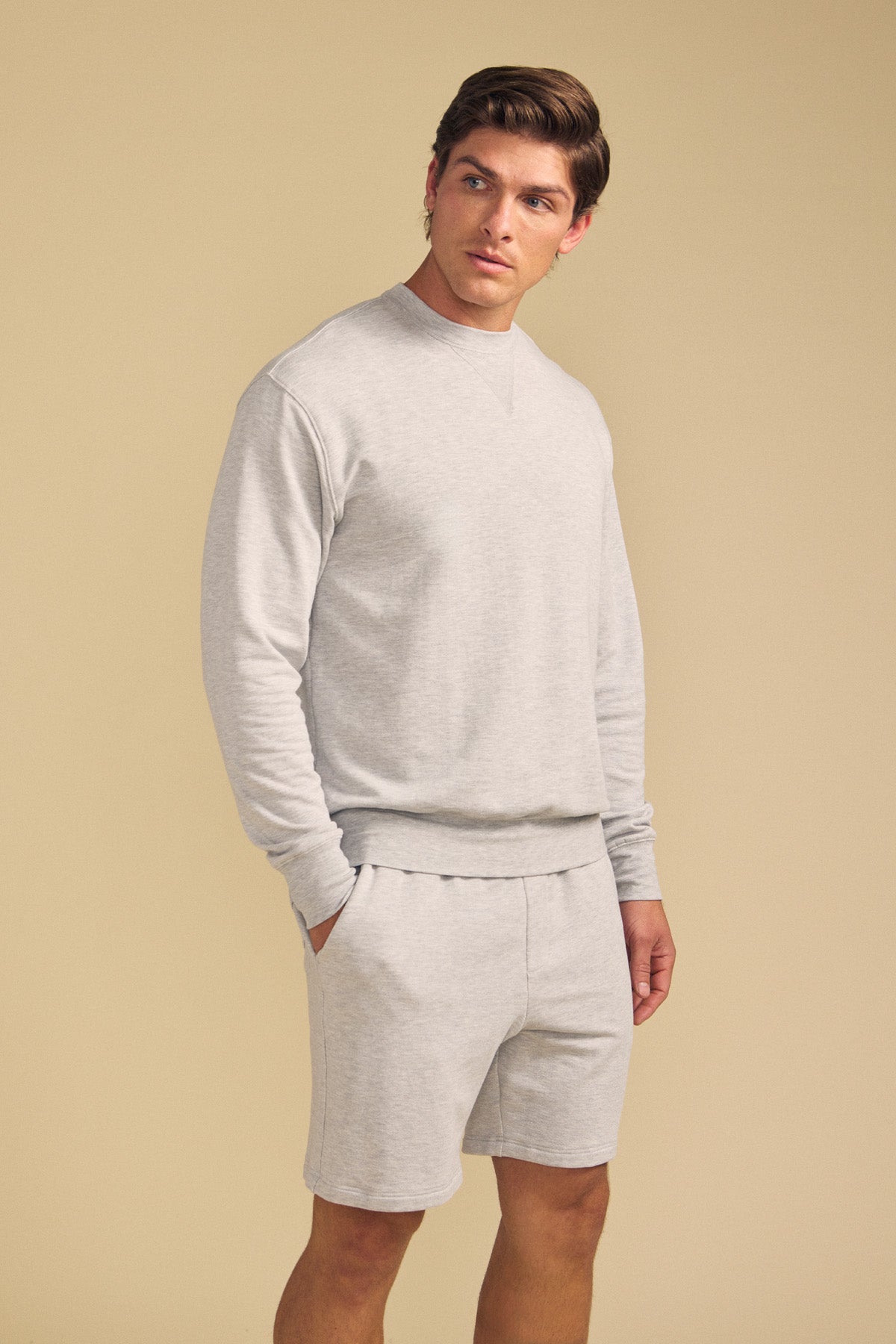 A man stands against a beige background wearing the OTIS SWEATSHIRT by Velvet by Graham & Spencer in light gray, paired with matching shorts, his left hand in his pocket.-41702161449153