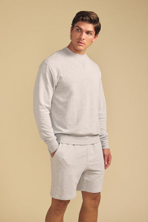 A man stands against a beige background wearing the OTIS SWEATSHIRT by Velvet by Graham & Spencer in light gray, paired with matching shorts, his left hand in his pocket.