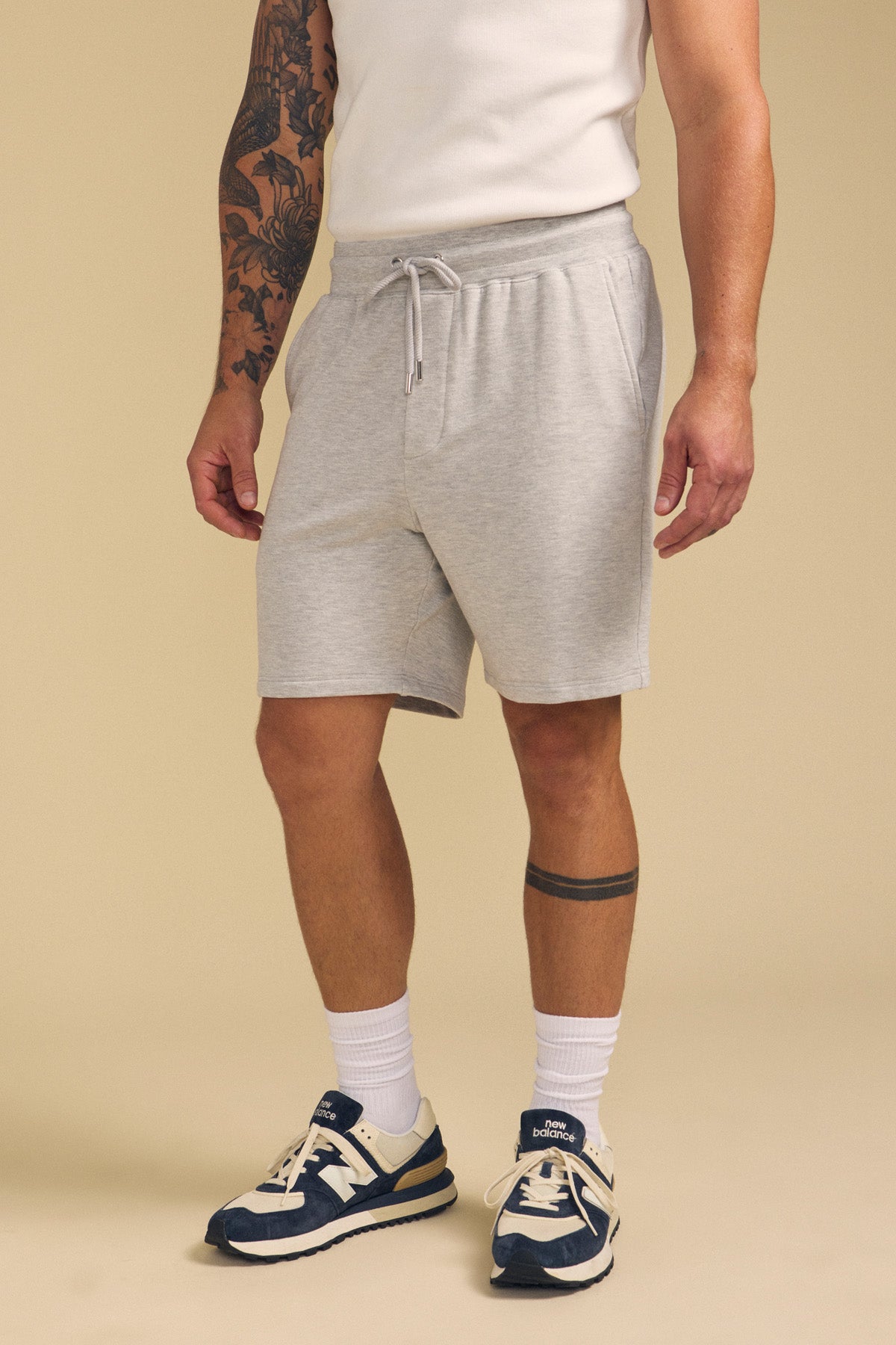 A person wearing a white sleeveless shirt, Velvet by Graham & Spencer QUENTIN SWEATSHORT in light gray, white crew socks, and navy sneakers stands against a plain beige background.-41702117572801