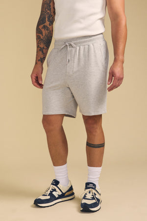 A person wearing a white sleeveless shirt, Velvet by Graham & Spencer QUENTIN SWEATSHORT in light gray, white crew socks, and navy sneakers stands against a plain beige background.