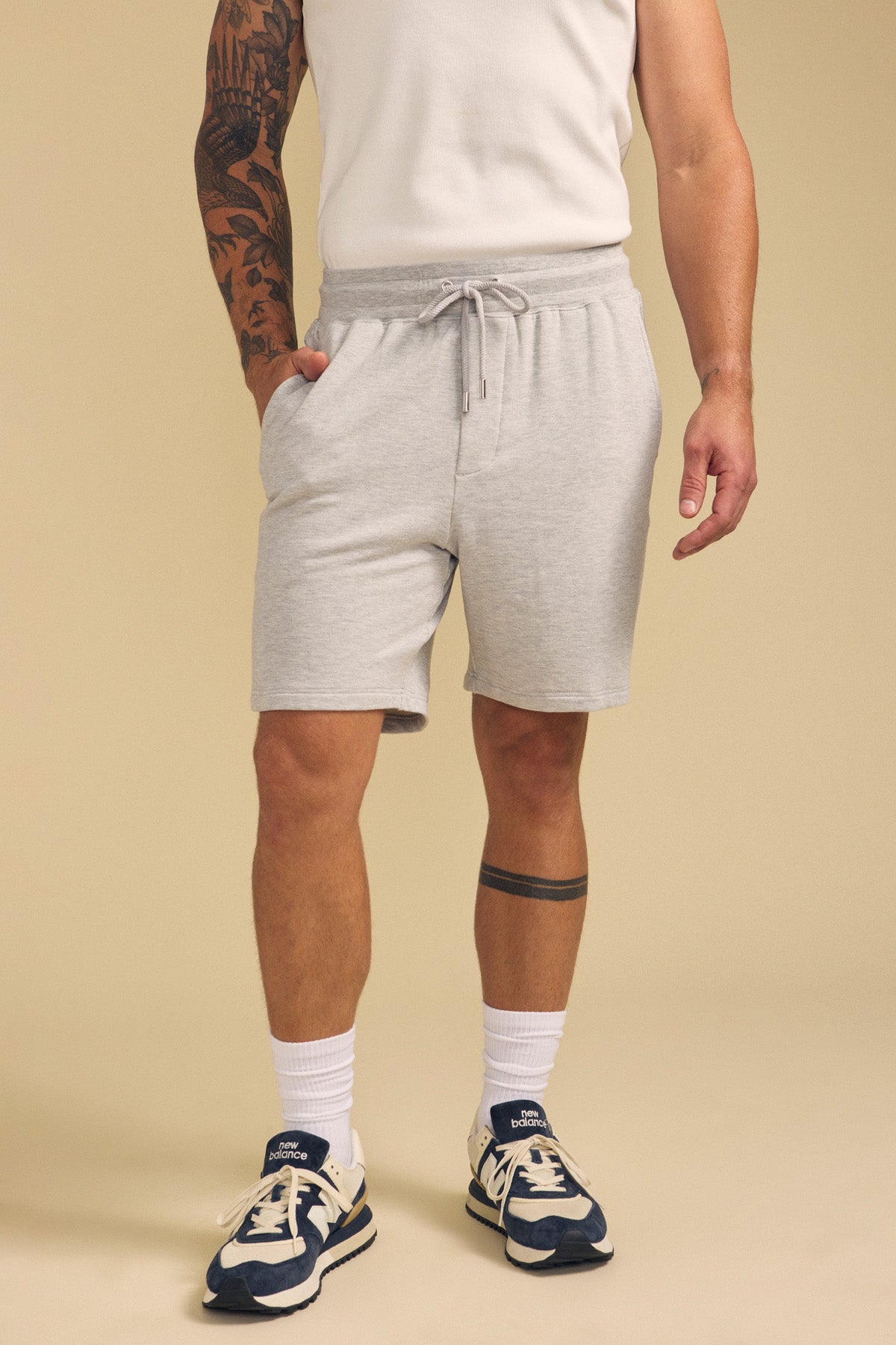 A person stands against a beige background wearing light gray Velvet by Graham & Spencer QUENTIN SWEATSHORTS, a white sleeveless shirt, white socks, and navy sneakers.