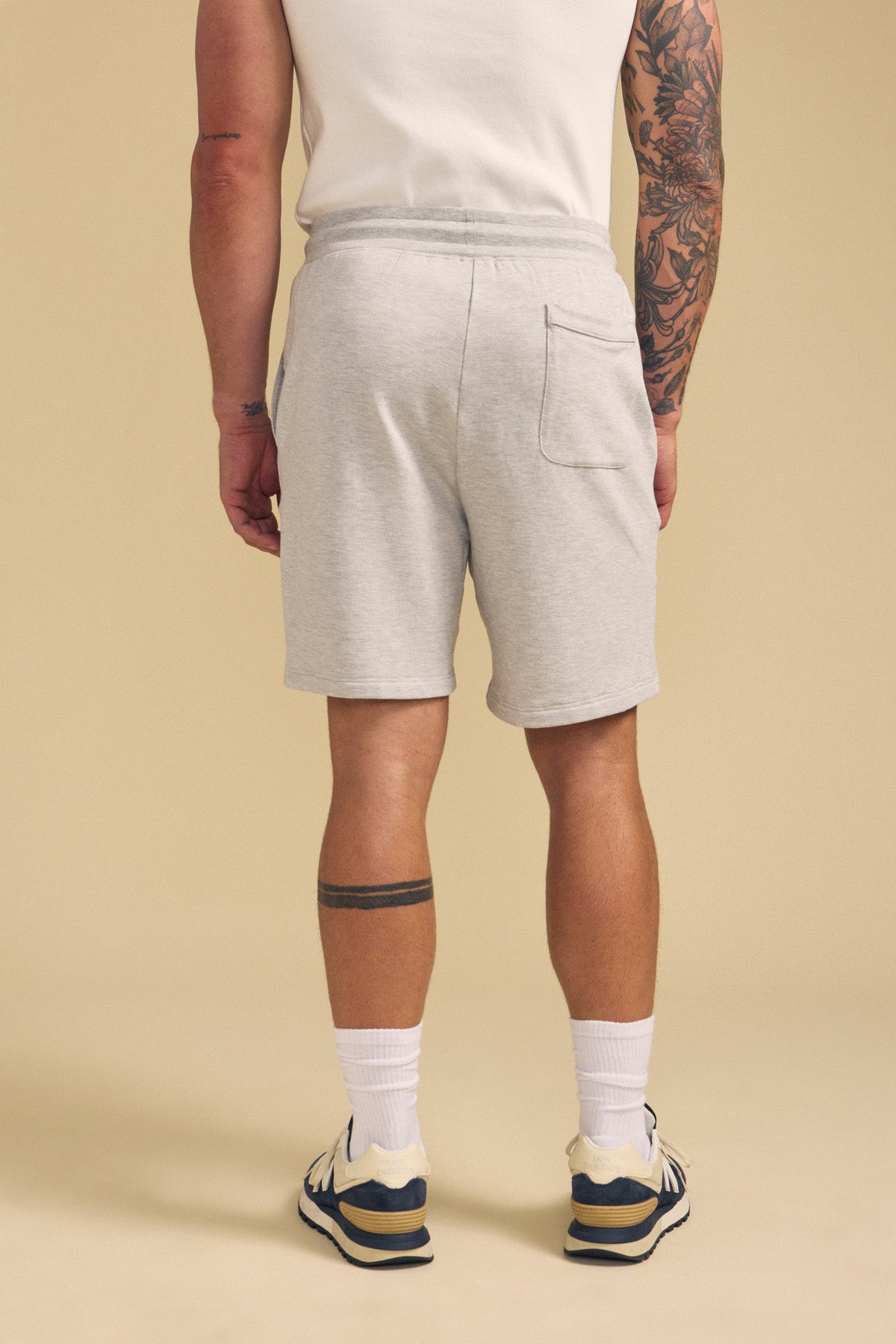 A man faces away, wearing Velvet by Graham & Spencer's QUENTIN SWEATSHORT in light gray, a white T-shirt, white socks, and sneakers. Tattoos are visible on his left arm and right calf.