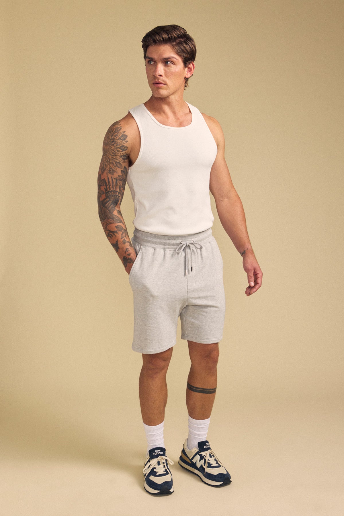 A man in a white tank top, Velvet by Graham & Spencer QUENTIN SWEATSHORT in light gray, white socks, and navy sneakers stands against a beige background.-41702117376193